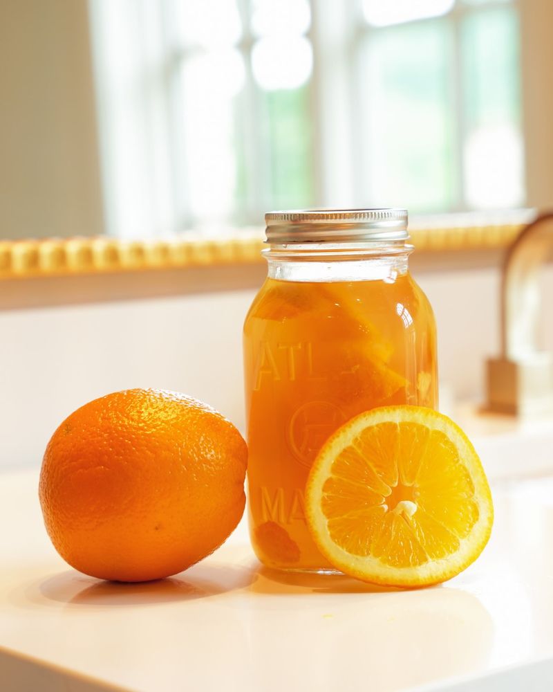 A glass jar of orange-infused liquid beside a whole orange and an orange slice on a kitchen counter